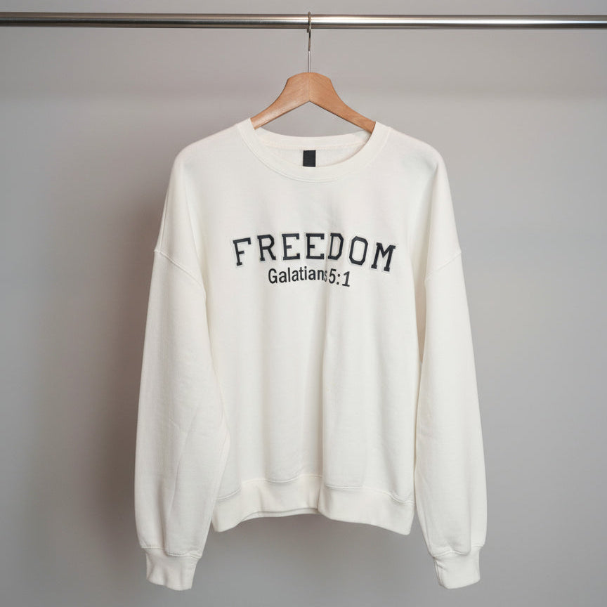 White sweatshirt with 'FREEDOM Galatians 5:1' text on a hanger against a tiled wall.