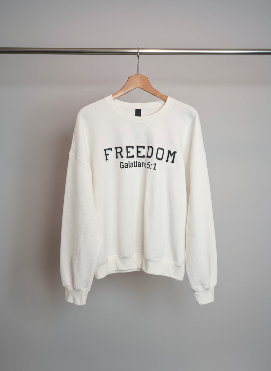 White sweatshirt with 'FREEDOM Galatians 5:1' text on a hanger against a tiled wall.